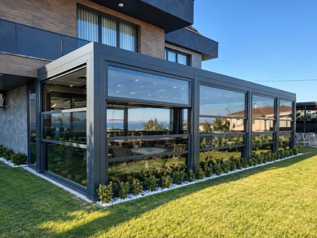 Giyotin cam sistemi, bahçeli modern villanın köşe camlı eklentisinde üst panelin yukarı kaldırılıp altında açık bir boşluk bıraktığı, gün ışığında dış mekândan görünüm
