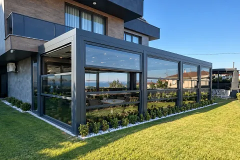 Giyotin cam sistemi, bahçeli modern villanın köşe camlı eklentisinde üst panelin yukarı kaldırılıp altında açık bir boşluk bıraktığı, gün ışığında dış mekândan görünüm